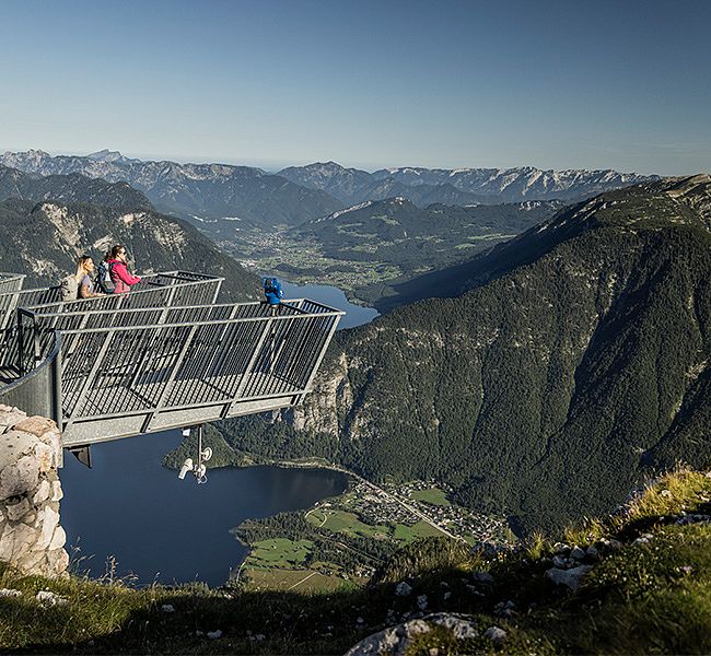 Metall-Aussichtsplattform ragt über eine Felsklippe. Drei Wanderer stehen am Geländer und schauen auf eine weite Berglandschaft mit einem See im Tal und hohen Bergen im Hintergrund.