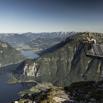 Person with a backpack stands alone on a metal observation deck high above a mountainous landscape with a lake in the valley and a clear sky.