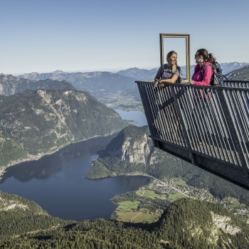 Two people with backpacks are standing on a metal observation deck high above a mountain landscape, with a lake in the background and a clear blue sky.