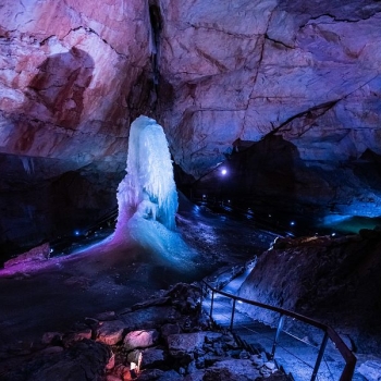 Interior view of an illuminated ice cave: In the center stands a large, column-shaped ice formation that extends from the ceiling to the floor. The cave walls are illuminated in reddish and purple hues. A secure walkway with a railing leads through the cave; the floor is wet and slippery in places. Small light sources along the path illuminate the space.