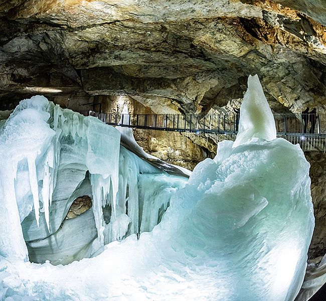 Blick in eine Eishöhle mit bläulich schimmernden Eiswänden und großen Eisformationen; ein Geländer verläuft entlang eines Gehwegs, während im Hintergrund Besucher die Höhle erkunden