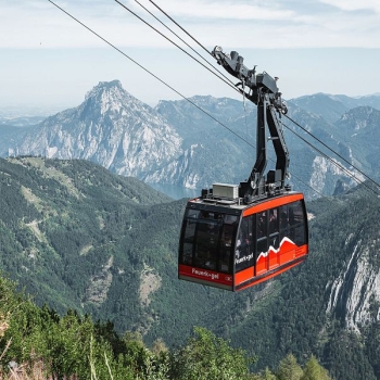 Moderne rote Seilbahnkabine fährt an Stahlseilen über eine bewaldete Berglandschaft mit steilen Felsgipfeln im Hintergrund.