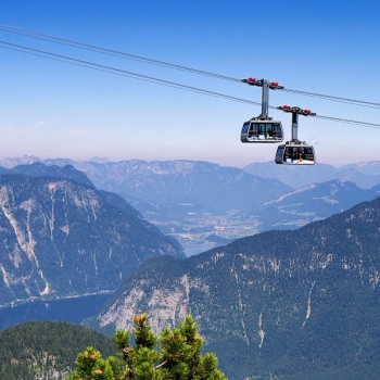 Two cable car gondolas glide over a mountainous landscape with forests, mountains, and a lake in the background.