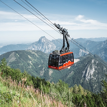 „Rot-schwarze Seilbahnkabine fährt über eine Berglandschaft mit Wald und Gipfeln; klare Himmelsszene im Hintergrund.“