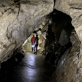 Zwei Wanderer in einer dunklen Höhle gehen durch eine enge Passage; raue Felswände umgeben sie, der Boden ist nass und reflektiert das Licht