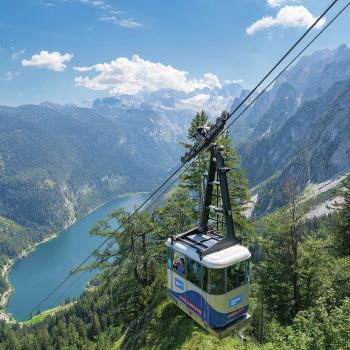 Seilbahn-Gondel in einer Alpenlandschaft über einem türkisblauen Bergsee; Drahtseile und steile Felswände im Hintergrund.