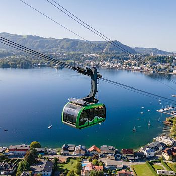 Eine grüne Seilbahngondel hängt an Drahtseilen über einem großen blauen See. Am Ufer erstreckt sich ein Ort mit Gebäuden, dahinter ragen Berge in den Himmel.