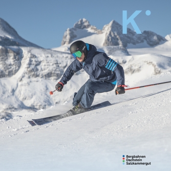 „Person in blauer Skibekleidung fährt in tiefer Kurve eine sonnige, breite Skipiste hinunter. Im Hintergrund ragen hohe, schneebedeckte Berggipfel in den blauen Himmel. Oben rechts ist ein großes, hellblaues ‚K‘ zu sehen, unten rechts der Schriftzug ‚Bergbahnen Dachstein Salzkammergut‘ mit drei farbigen Punkten davor.“